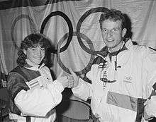 A woman with shoulder length, curly hair and a man with short, curly hair bump fists.  They are both in white coats with dark shoulders