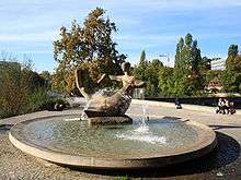 Circular fountain surrounded by pavement. The fountain's centre is a sculpture of a pair of abstract human figures.