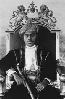 A black-and-white photograph of a man with a dark moustache wearing a turban and a dark jacket and sitting on a throne topped by two metal lions