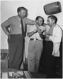 Three men talking. The one on the left is wearing a tie and leans against a wall. He stands head and shoulders above the other two. The one in the centre is smiling, and wearing an open-necked shirt. The one on the right wears a shirt and lab coat. All three have photo ID passes.